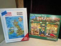 Two boxed 1000-piece puzzles side by side. Left puzzle is a UK ocean map by Peoples Friend, right puzzle is Vintage Shop scene.
