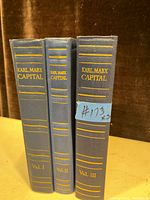 Three blue hardcover books titled 'Karl Marx Capital' volumes I, II, and III shown standing vertical. Volume III has a blue sticker label. All have gold text and lines on spine.