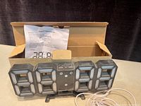 3D digital alarm clock with three buttons on top, four large 8-shaped LED digits, white USB cable, and instruction paper behind, with original box open in background.