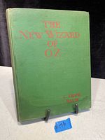 Green hardcover book front cover with red text 'THE NEW WIZARD OF OZ' by L. Frank Baum. Shows some wear and slight fading.
