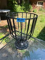 Black iron flower pot holder placed on a glass outdoor table in a garden setting, showing vertical bars, scrollwork details, and circular base.