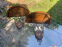 Pair of semi-circular wooden shelves with decorative iron scrollwork brackets placed on a glass table outside on grass.