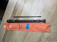 Top-down view of dark wood soprano flute, orange soft case, and black cleaning rod on wood floor