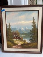 Full view of the framed oil painting depicting a mountain landscape with pine trees, cabin, lake and walking figures.