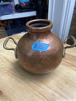 Photo showing a round vintage solid copper vase with two cast brass handles on either side, placed on a wooden surface, with visible dings and a blue tape label marked #43.