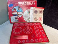 Photo of vintage Spirograph set showing red plastic base with multiple gears, original box, and instructional booklet.