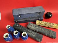 Photo showing four small blue vases, three intricately carved stone mortars with gold and gilt decorations, and two carrying cases. A small black fruit-shaped ceramic object and a green apple for size reference included.