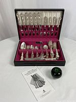 Open black wooden presentation box displaying silver plated flatware set with burgundy interior, instruction booklet, and green glass apple