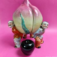 Front view of colorful porcelain figurine showing three men holding a large peach with a black apple placed in front for size reference.