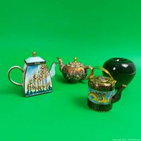 Three miniature teapots shown with a green apple used for size reference. Teapots vary in shape, color, and enamel detailing.