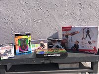 Overview of all boxed games and toys in the lot placed on a outdoor black bench against white wall.