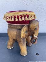 Side view of hand-carved wooden elephant stool with red velvet cushion featuring tassel fringe