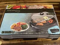 The original box of the Kitchen Living electric indoor grill showing food grilled on the unit and brand information. Box is worn and coming apart.