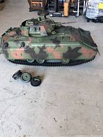 Side view of large camouflaged G.I. Joe tank toy with broken wheels and extra wheels beside it.