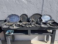 Wide view of entire lot displayed on black bench showing all silver plated items including trays, teapots, cups, and small dishes.