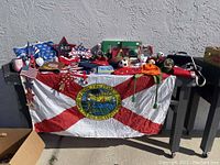 Table covered with a large Florida state flag displaying multiple miscellaneous items including patriotic stars, holiday decorations, a soccer ball, plush toys, and other small items.