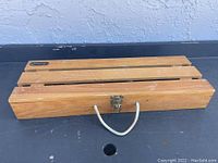 Closed view of wooden carrying case with horizontal slats, metal latch, and rope handle on black surface