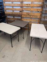 Three folding tables shown: two white rectangular tables and one gray square card table, all with black metal legs, positioned together in front of a wooden pallet wall.