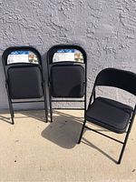 Three black metal folding chairs with padded fabric seats and backs, set against wall outdoors under sunlight