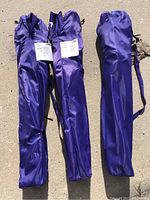 Three purple foldable chairs packed in matching purple carrying bags, each bag tall and slim with a carrying strap and a tag attached.