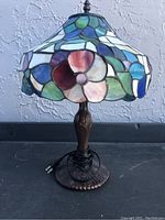 Full view of Tiffany style lamp showing stained glass shade with flower and leaf pattern, metal base, electrical cord and plug.