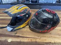 Two motorcycle helmets side by side on wooden surface: one off-road style in yellow and black with flames, one black full-face with red accents and clear visor