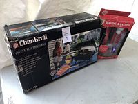 Photo showing Char-Broil Deluxe Electric Grill boxed unit alongside a red roasting and basting kit in packaging, placed on a light surface.