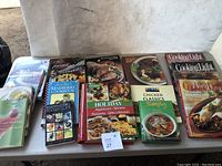 Photo shows an assortment of cooking books arranged side by side on a table, titles visible, colors vary.