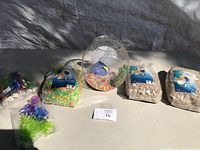 Fish bowl with colorful gravel inside surrounded by three bags of aquarium substrate and two decorative aquatic plants on a table outdoors.