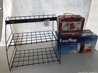 Photo of black metal wire coffee pod caddy with two shelves alongside vintage ABC lunchbox and boxed EasyNap napkin dispenser on white background