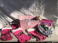Lot sundry Christmas decorations on a table under daylight including pink stockings, ornaments in basket and bag, boxed decoration, and silver tinsel garland