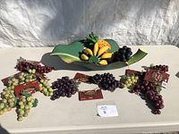 Overview of lot with green leaf bowl holding bananas and pineapple surrounded by five bunches of artificial grapes on a table.