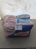 Photo showing two large sealed packs of Tena Woman disposable underwear diapers, with pink and blue packaging. One pack is size XL and the other large size, designed for bladder support.