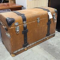 Front view of vintage automotive storage trunk showing brown cover with black leather corner protectors, metal locks, and straps.