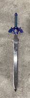 Full length image showing the cosplay sword lying flat on carpet, highlighting the silver blade and blue handle with wing-like crossguard.