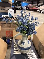 Ceramic pitcher vase with artificial blue flowers in warehouse setting on boxes