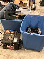 Wide view showing Dremel table saw on floor next to blue storage bins containing various tools like Little Giant leg leveler, power drill, and accessories.