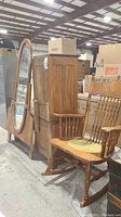 Photo showing vintage wooden dresser with attached large oval mirror leaning against it and a wooden rocking chair beside them in a warehouse setting.