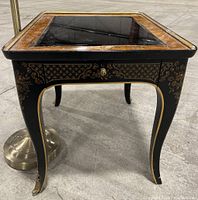 Full view of Drexel side table showing black lacquer finish, gold floral and geometric designs, glass top with wood border, minor water damage and scratches on top.