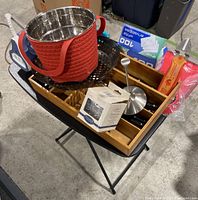 Top view of the black foldable table displaying a red silicone collapsible strainer, metal steamer basket, bamboo drawer organizer with knives and utensil tool, and office supplies.
