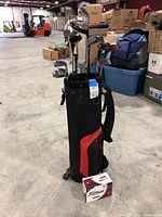 Photo showing black and red golf bag standing with assorted golf clubs inside, new sealed Titleist golf ball box placed in front.