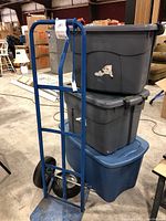 Photo showing blue Marathon hand truck with two tires, one detached, and stacked plastic storage totes in gray and blue colors.