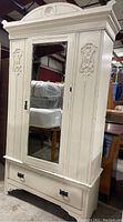 Full view of the antique armoire showing the clock with mirror front door and base drawer, carved side panels, and detailed crown molding.
