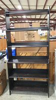 Full front view of black wooden shelving unit showing five shelves and overall form in warehouse setting.