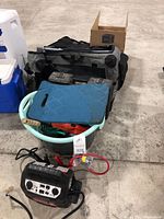 A bucket containing various tools, a blue foam kneeling pad, a soft pet carrier bag, and a portable power pack with cables on the floor.