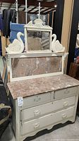 Photo showing front view of vintage white painted dresser with pinkish marble top, back panel with marble inset, and framed mirror with carved swan figures on top.