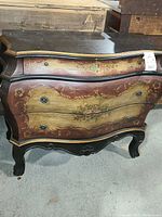 Front view of the vintage wooden dresser showing curved drawers with decorative floral paint and vintage knobs. Visible signs of wear and paint distress.
