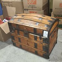 Front view of the vintage wooden trunk showing domed top, metal straps, lock latch, and visible wear including water damage and scratches.