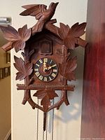Front of wooden cuckoo clock showing bird on top, carved leaves around dial, and hanging pendulum chain.