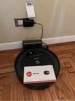 Hoover Quest 1000 robotic vacuum on wooden floor with power adapter plugged in and instruction manual resting on it.
